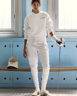 A person in white athletic gear stands indoors holding a fencing sword, with a fencing helmet on a wooden bench nearby. They wear sneakers, a sweatshirt, and sweatpants, against a blue wall and windows.