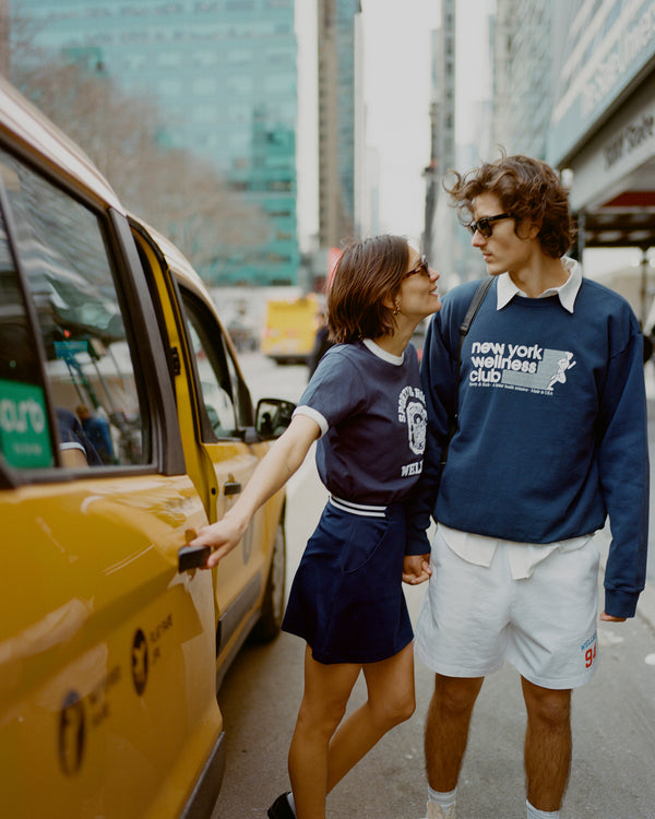 A couple stands by a yellow taxi, holding hands. She wears the Golf Logo Kelly Skirt - Navy/White, its high-quality fabric draping elegantly as they pause amidst the citys bustle.