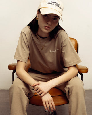 Young woman wearing the Drink More Water T-Shirt in Elephant/White, made from USA-grown cotton, sits on a brown chair leaning forward with hands clasped.