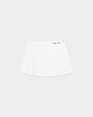 A woman wearing glasses, beige shoes, a navy V-neck sweater, and the Abigail Signature Logo Abigail Skirt in Off White stands against a white wall.