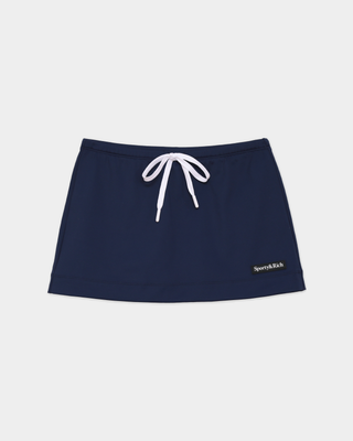 A woman stands against a white background wearing a red, white, and blue striped rugby shirt, tan shoes, and the Azalee Serif Logo Tie Skirt in Dark Navy featuring an adjustable waist and built-in shorts.