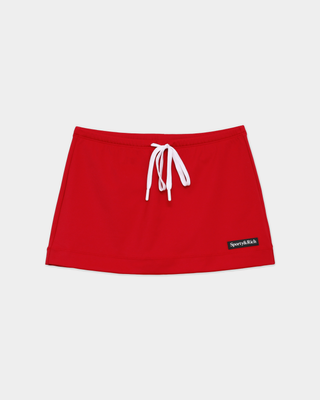 A woman is wearing the Azalee Serif Logo Tie Skirt in Ruby—a red skirt with an adjustable waist and built-in shorts—styled with a red collared sweatshirt, tan shoes, and a watch, as she stands against a white wall.