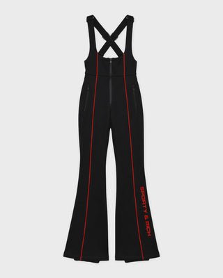A person wears a black Divy Bold Logo Ski Jumpsuit with red piping, a red headband, and yellow sunglasses.