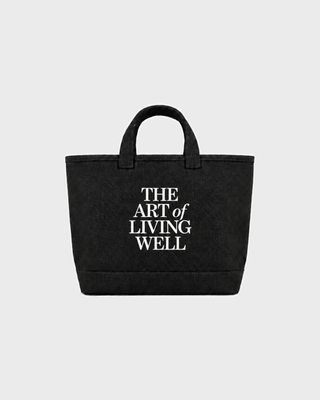 A black canvas tote bag with two top handles and bold white text on the front reading 'THE ART of LIVING WELL', shown against a light gray background.