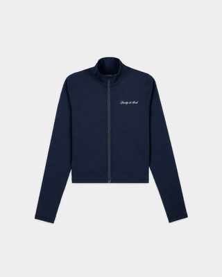 A woman in glasses wears the Freya Signature Logo Sports Jacket in Dark Navy, crafted from technical stretch fabric, paired with light gray sweatpants, standing against a plain white wall.