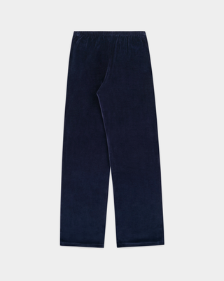 A woman stands against a plain white background wearing the Benedicte S Logo Velour Jogger in Dark Navy/White—wide leg velour track pants with an adjustable drawstring waistband—paired with beige shoes.
