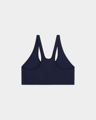 A woman stands against a plain white wall in the Billie Signature Logo Sports Bra - Dark Navy, arms at her sides and ready for high-impact workouts, supported by its racerback design.