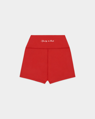 A woman stands against a plain white background, her hair in braids and wearing sneakers, dressed in a red sports bra and the Blaire Signature Logo Mini Biker Short in Ruby. The high-waist fit and super soft fabric of these shorts ensure comfort while she moves.