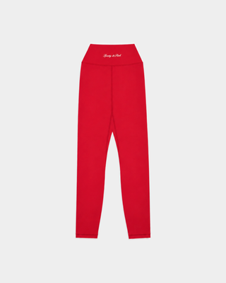 A woman stands against a white background wearing the Bonnie Signature Logo Legging in Ruby, paired with a matching sports bra and yellow and black sneakers. These opaque workout leggings deliver a supportive fit for any fitness routine.