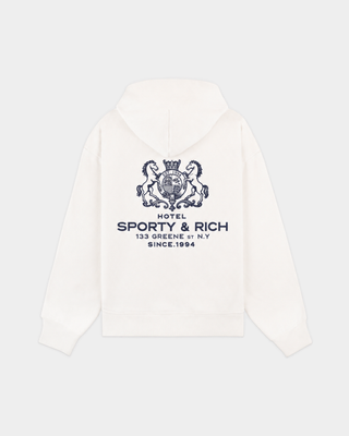 Woman in the Elaine SR Hotel Hoodie - Off White and cream sweatpants stands against a white wall, holding her hood with one hand.