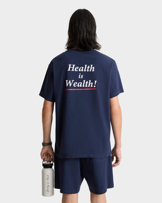Person wearing the Health is Wealth T-shirt in navy, garment-dyed with “Health is Wealth!” on the back, paired with shorts and holding a water bottle. Made in the USA for comfort and quality.