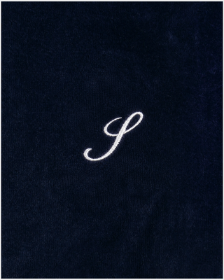 A woman stands against a plain white background wearing the Benedicte S Logo Velour Jogger in Dark Navy/White—wide leg velour track pants with an adjustable drawstring waistband—paired with beige shoes.