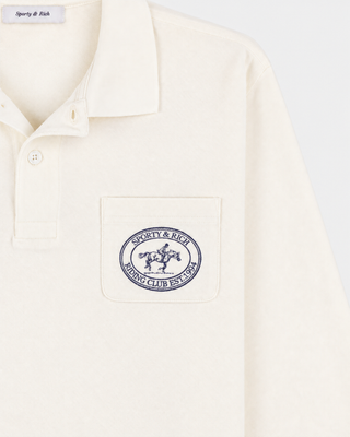 Introducing the Perry Riding Club Longsleeve Pique Polo in Off White—a cream-colored, oversized long-sleeve polo shirt made from 100% cotton piqué. It features a small chest pocket detailed with a blue bear and text logo patch, combining comfort with playful charm.