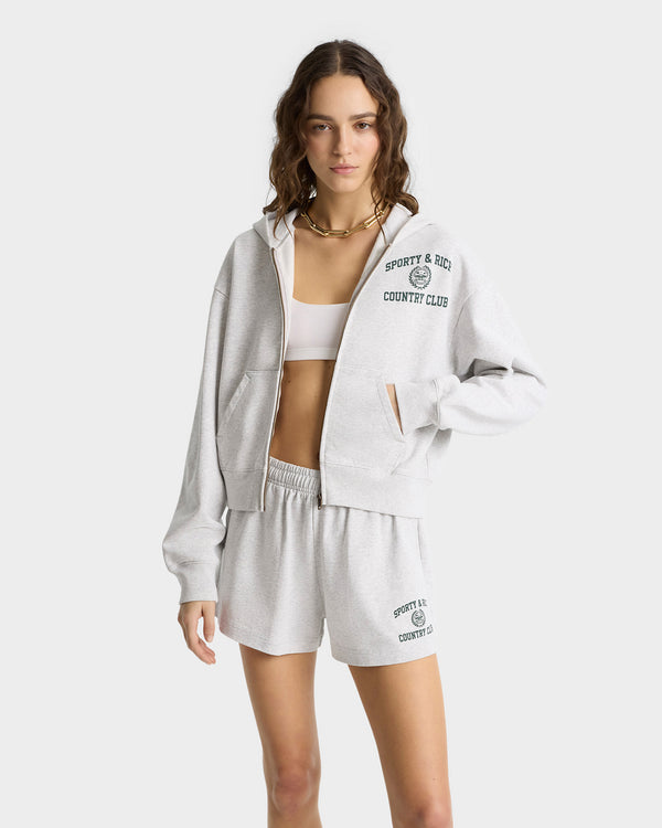 A person in an oversized gray hoodie and shorts, with a white sports bra, poses against a plain background. The Varsity Crest Cropped Zip Hoodie in Heather Gray/Forest gives an effortlessly chic look, showcasing the quality craftsmanship typical of garments made in Portugal.