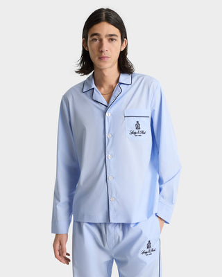 A person wearing Vendome Pyjama Pants in Ciel/Navy—light blue cotton trousers with dark piping, elastic waistband, and embroidered logo—stands against a plain background.