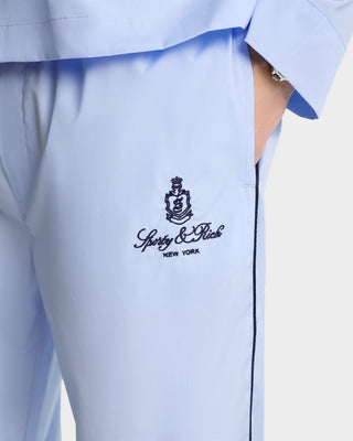 Person wearing Vendome Pyjama Pants - Ciel/Navy, light blue cotton trousers with Sporty & Rich New York embroidered on the pocket, elastic waistband, hand in pocket. Made in Portugal.