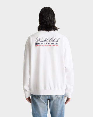 Person wearing the Made In USA Crewneck - White/Navy/Fire Red, a white garment-dyed sweatshirt with Health Club SPORTY & RICH text on the back, paired with light blue jeans.