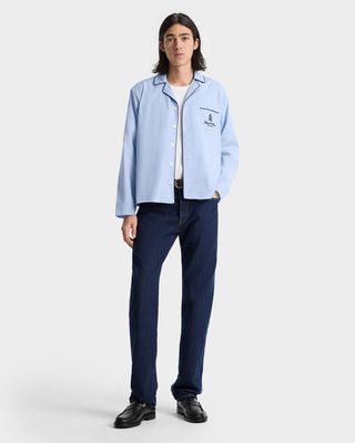 A man wears the Vendome Pyjama Shirt - Ciel/Navy with an embroidered logo, paired with dark jeans and black shoes, standing against a plain background.