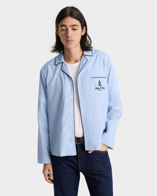 A person wears the Vendome Pyjama Shirt - Ciel/Navy, a light blue cotton shirt with dark piping and an embroidered logo, paired with dark jeans, standing against a plain background.