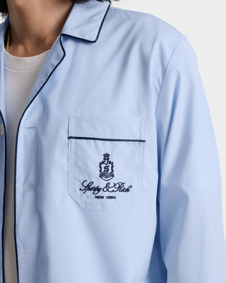 A person wearing the Vendome Pyjama Shirt - Ciel/Navy, a light blue cotton shirt with navy-trimmed pocket, embroidered crest, Sporty & Rich NEW YORK logo, made in Portugal.