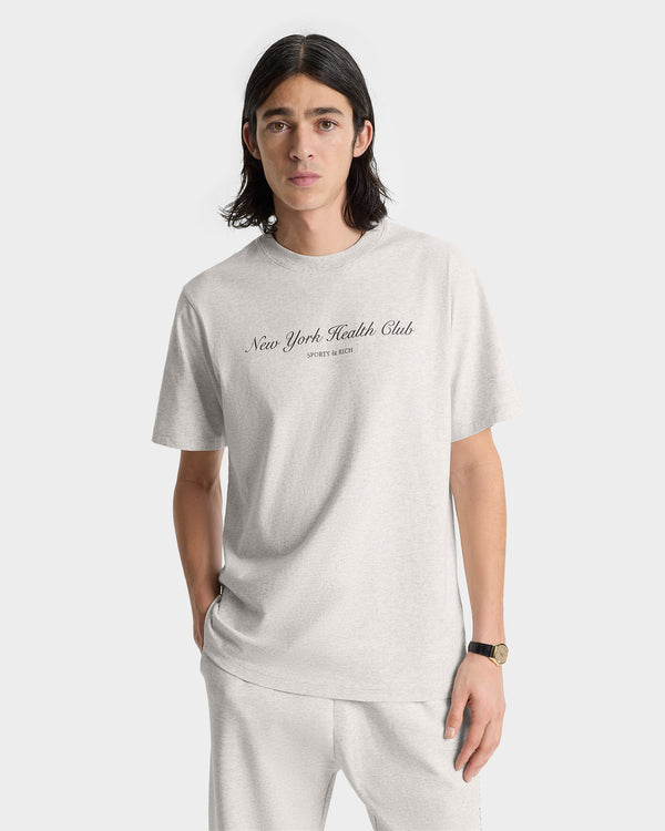 Man in the NY Health Club T-Shirt - Heather Gray/Navy, made from 100% shrink-free cotton and crafted in Portugal, stands against a white background. New York Health Club is printed on the shirt for lasting comfort and style.