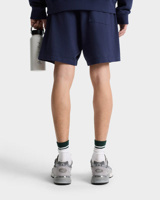 Person wearing Royal Club Gym Short - Navy/White with an elastic waistband and sneakers, holding a water bottle, shown from behind against a plain background.