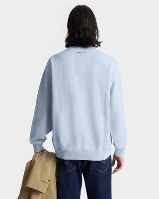 Person with shoulder-length hair facing away, wearing the Wellness Club Flocked Crewneck in Ciel/Dark Navy (100% cotton), and holding a tan coat.