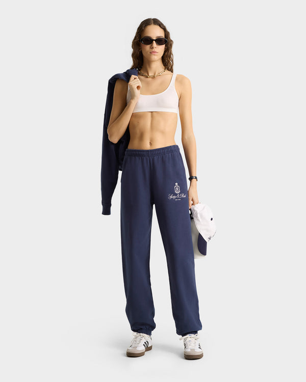 Woman in sunglasses, white crop top, Vendome Sweatpant - Navy/White made in Portugal, sneakers, holding a navy jacket and white cap.
