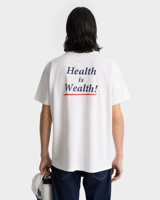 Person wearing the Health Is Wealth T-Shirt - White/Navy/Fire Red, an oversized white tee made in the USA with Health is Wealth! printed on the back in blue and red letters.