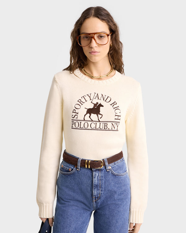 A glasses-wearing individual dons the Sporty & Rich Polo Club Knitted Sweater in Cream/Chocolate with an embroidered logo, pairing it with blue jeans and a clutch.