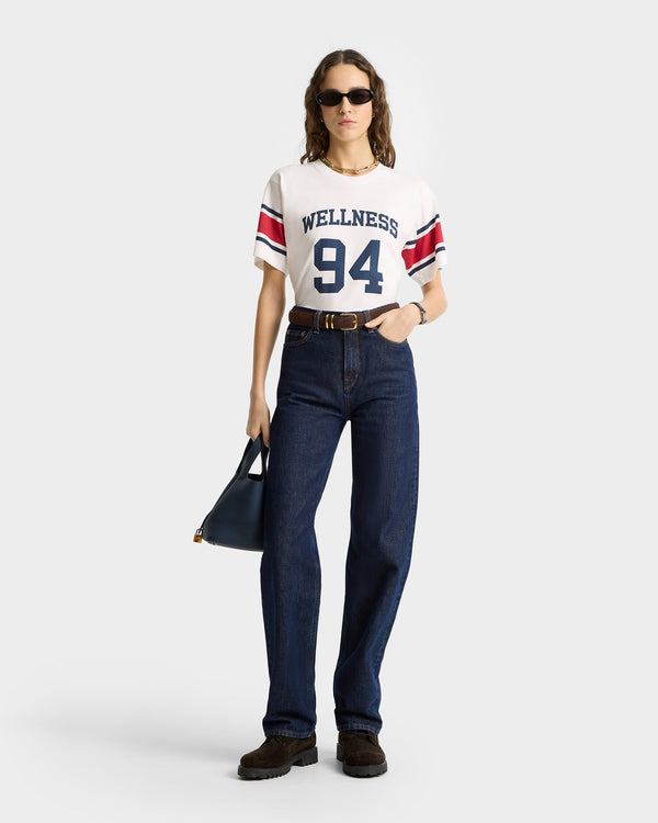 A person in sunglasses wears a Wellness 94 Rugby Tee in white, ruby, and navy with a vintage look, blue jeans, and holds a black handbag.