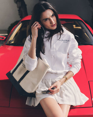 A woman in a white outfit sits on a red car, holding the NY Health Club Tote Bag - Natural/Navy and her phone, listening to music with earphones.