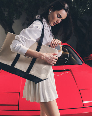 A woman in white stands by a red sports car with the NY Health Club Tote Bag - Natural/Navy, listening to earphones.