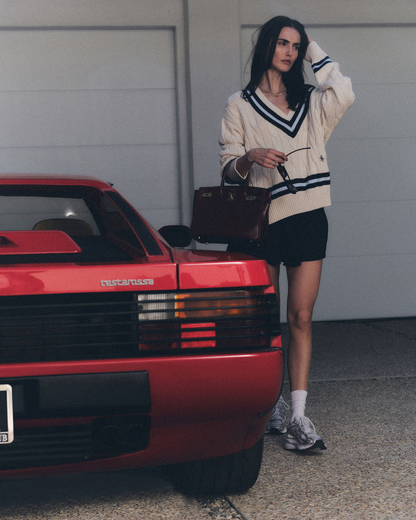 A woman in a cream sweater and SRC Logo Abigail Skirt - Navy/White stands by a red sports car, holding a brown handbag.