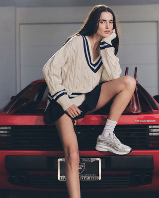 A woman in a sweater and sneakers, wearing the SRC Logo Abigail Skirt in Navy/White, sits on the back of a red sports car, posing with one hand on her face.