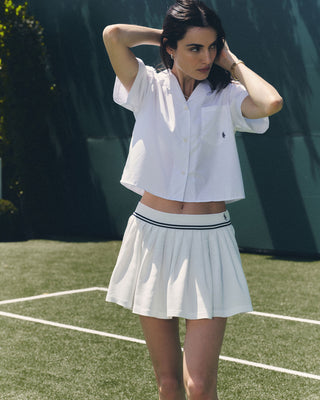 A woman wearing a white short-sleeve shirt and the SRC Logo Abigail Skirt in White/Navy stands on a tennis court, adjusting her hair.