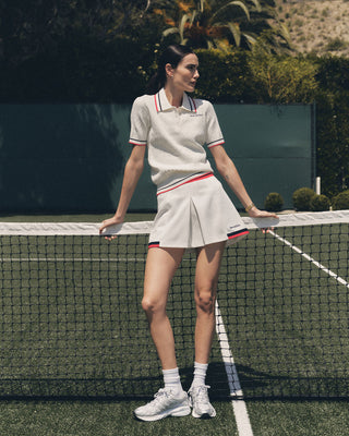 A woman in a white Serif Logo Mila Skirt (White/Navy/Fire Red) leans against an outdoor tennis court net.