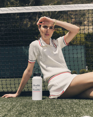A woman in tennis attire sits by the court net, shading her eyes, wearing the Serif Logo Mini Cable Polo in White/Navy/Ruby, and holding a water bottle labeled Health is Wealth!.
