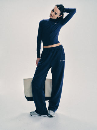 Woman wearing the Syracuse Embroidered Wide sweatpant in navy/white and sneakers stands holding a large tote bag with an embroidered logo, posing with one hand behind her neck.