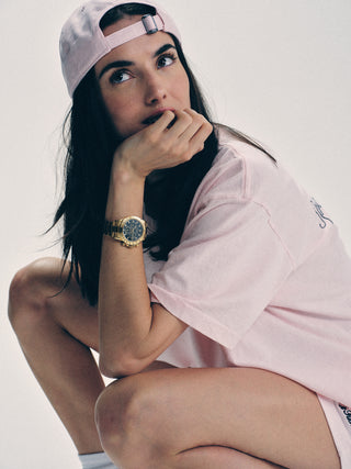 A woman wearing the Syracuse Hat - Pink Dolphin, with an adjustable closure, sits thoughtfully in a pink cotton twill shirt and wristwatch, resting her chin on her hand.