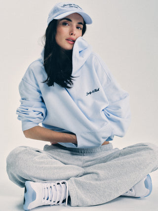 A woman in a light blue hoodie, gray sweatpants, and the Syracuse Hat - Ciel with an adjustable cotton twill design sits cross-legged on the floor against a white background.