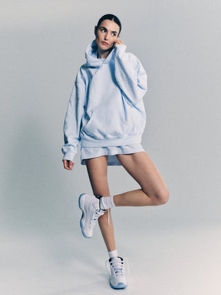 Woman in the Syracuse Embroidered Hoodie - Ciel/Navy poses thoughtfully, wearing a mini skirt and sneakers with one leg raised.