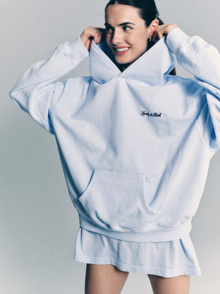 Smiling woman in the Syracuse Embroidered Hoodie - Ciel/Navy and a short skirt, holding the hood with both hands.