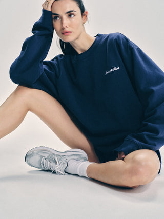 A woman wearing the Syracuse Embroidered Crewneck in Navy/White, made from USA-grown cotton, and sneakers sits on the floor with one hand resting on her head.