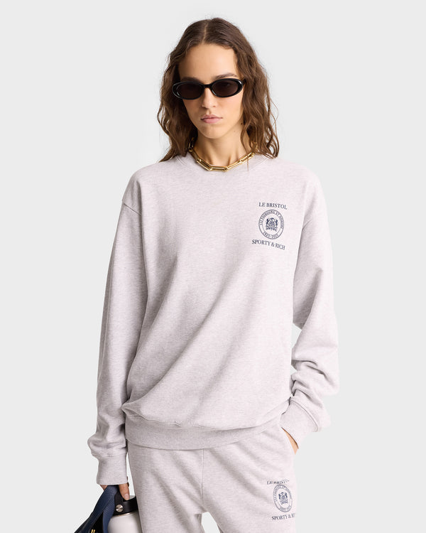 Woman in the Crest Seal 100th Crewneck – Heather Gray/Navy, oversized fit for a relaxed look, paired with sweatpants. Features sporty Le Bristol Sporty & Rich logos, sunglasses, and a necklace for effortless style.