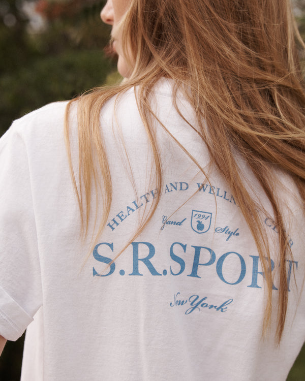 A woman with long hair wears a white Wimbledon T-Shirt - White/Washed Hydrangea, featuring blue text on the back, outdoors.