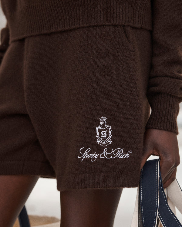 Close-up of a person wearing Vendome Cashmere Short in Chocolate/White with an elastic waistband and embroidered Sporty & Rich logo, holding a cream and navy bag.
