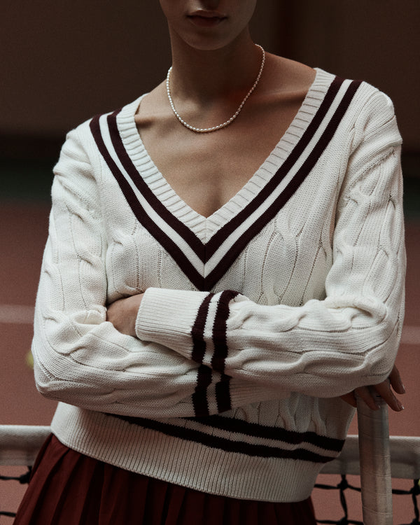 Woman wearing the SRC Cableknit V-Neck Sweater in Off White/Merlot, featuring dark stripes, paired with a pearl necklace and standing with arms crossed.