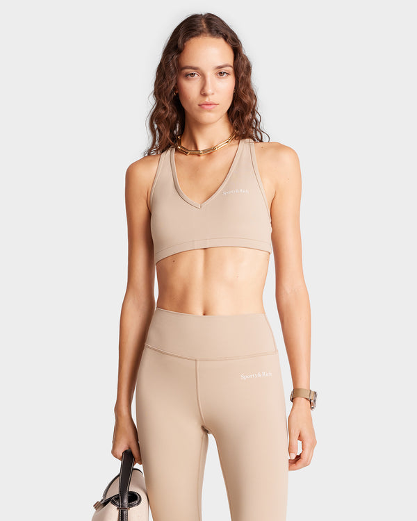 A woman in luxury activewear, featuring the elegant Serif Logo V Neck Sports Bra in Elephant/White, holds a yoga mat. She stands confidently against a simple background, exuding an effortless blend of style and fitness.