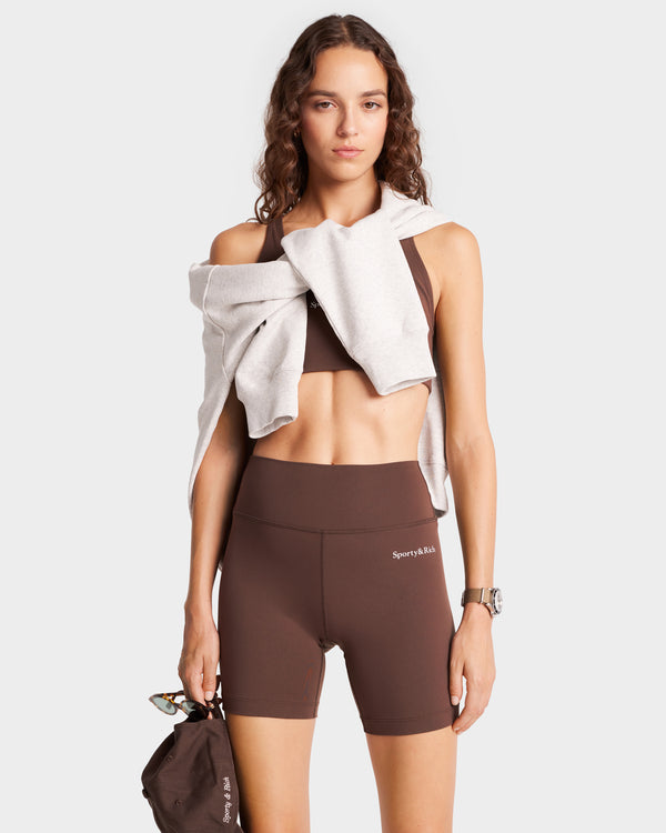 A woman exudes effortless style in Serif Logo Biker Shorts - Chocolate/White, with a sweatshirt draped over her shoulders. She holds a brown bag and wears a watch, perfectly blending fashion and function.